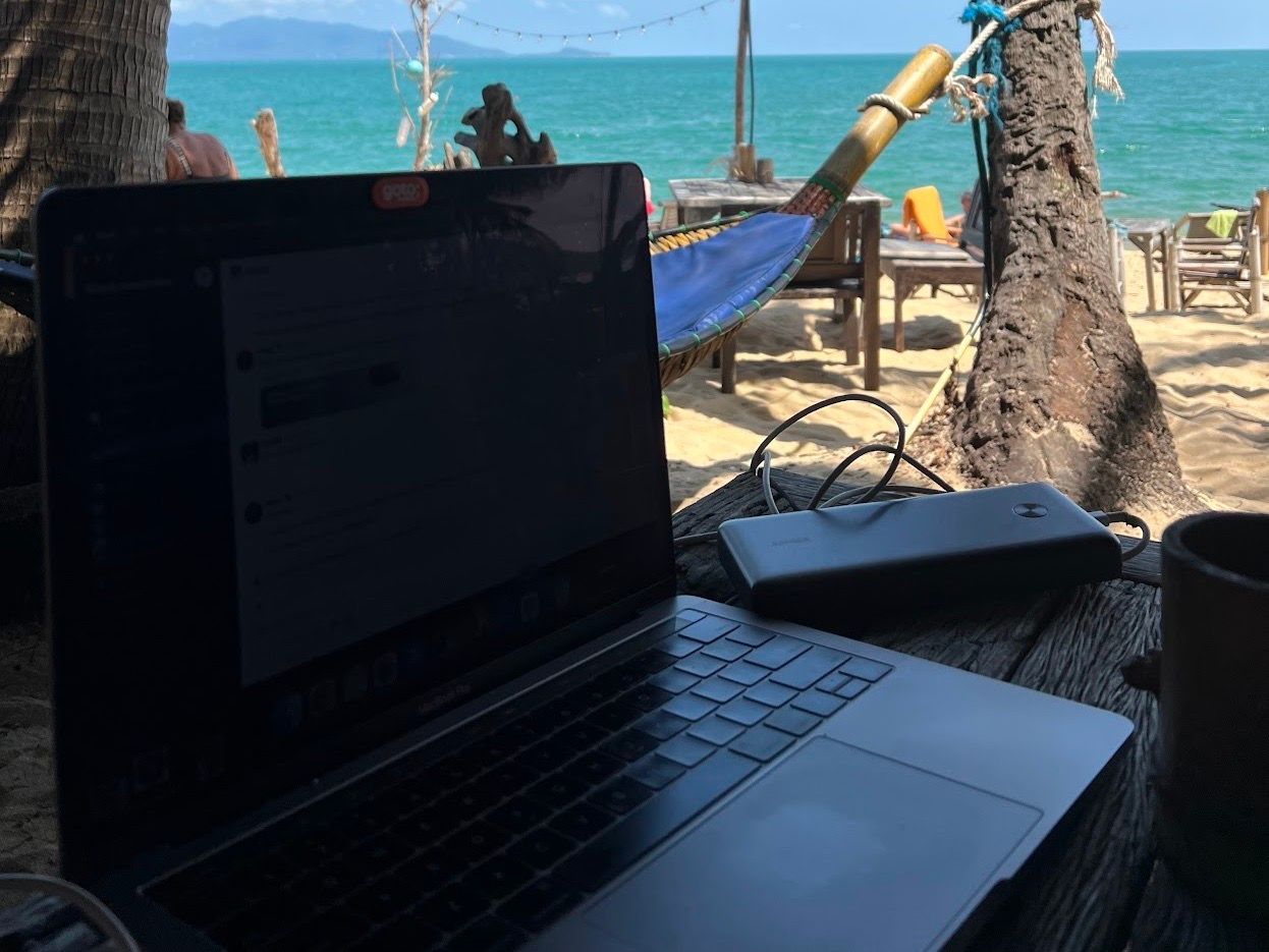 Apple MacBook laptop with an Anker powerbank on a wooden table on the beach. Ocean in the background with a tree and a hammock. Hua Kati Cafe, Koh Samui, Thailand