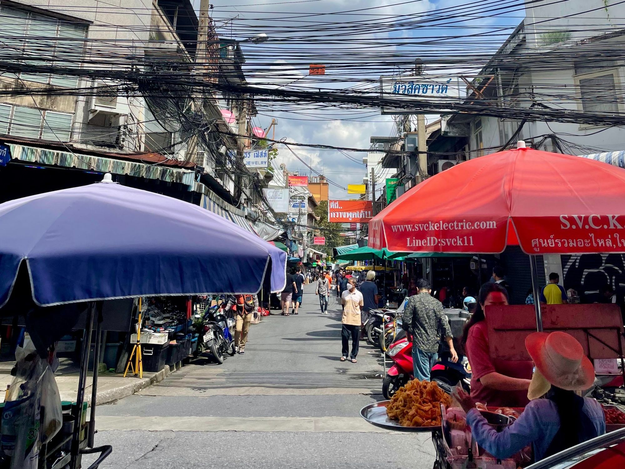 Bangkok, Thailand - A busy street with stalls, people wearing mouth masks, and electric wires above the ground.