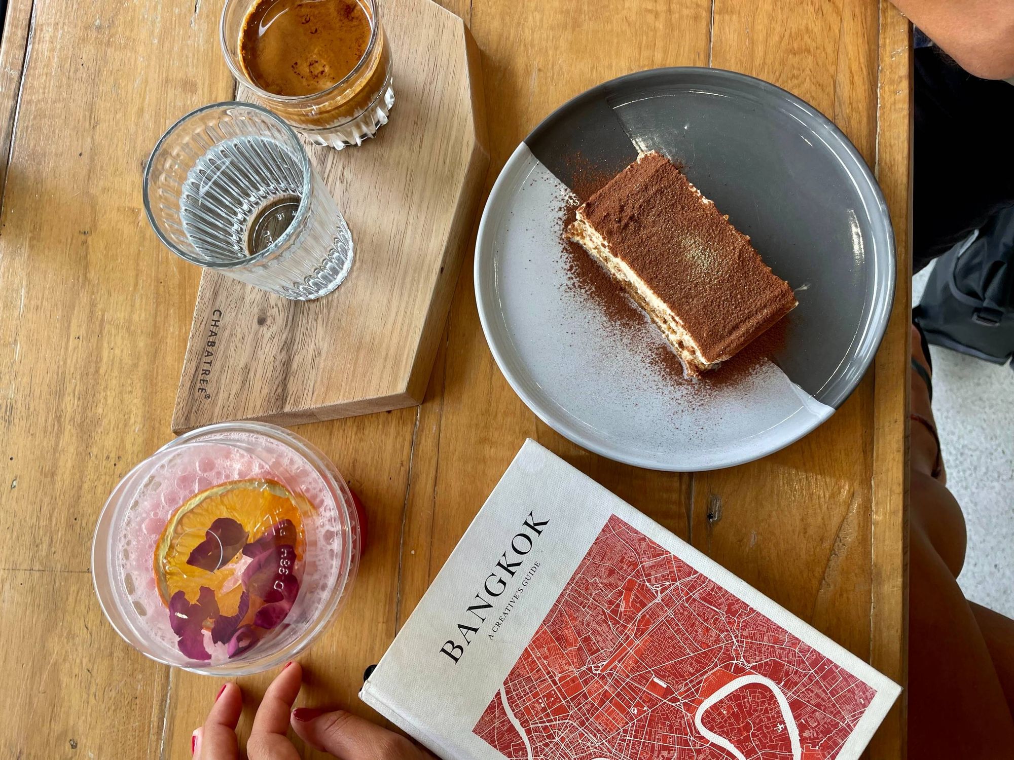 A new cafe, Varen, is located in Bangkok, Thailand. Wooden table with a pink drink with an orange and a flower. A coffee with a glass of water next to it./ A creatives guide about Bangkok and a tiramisu topped with gold glitter.