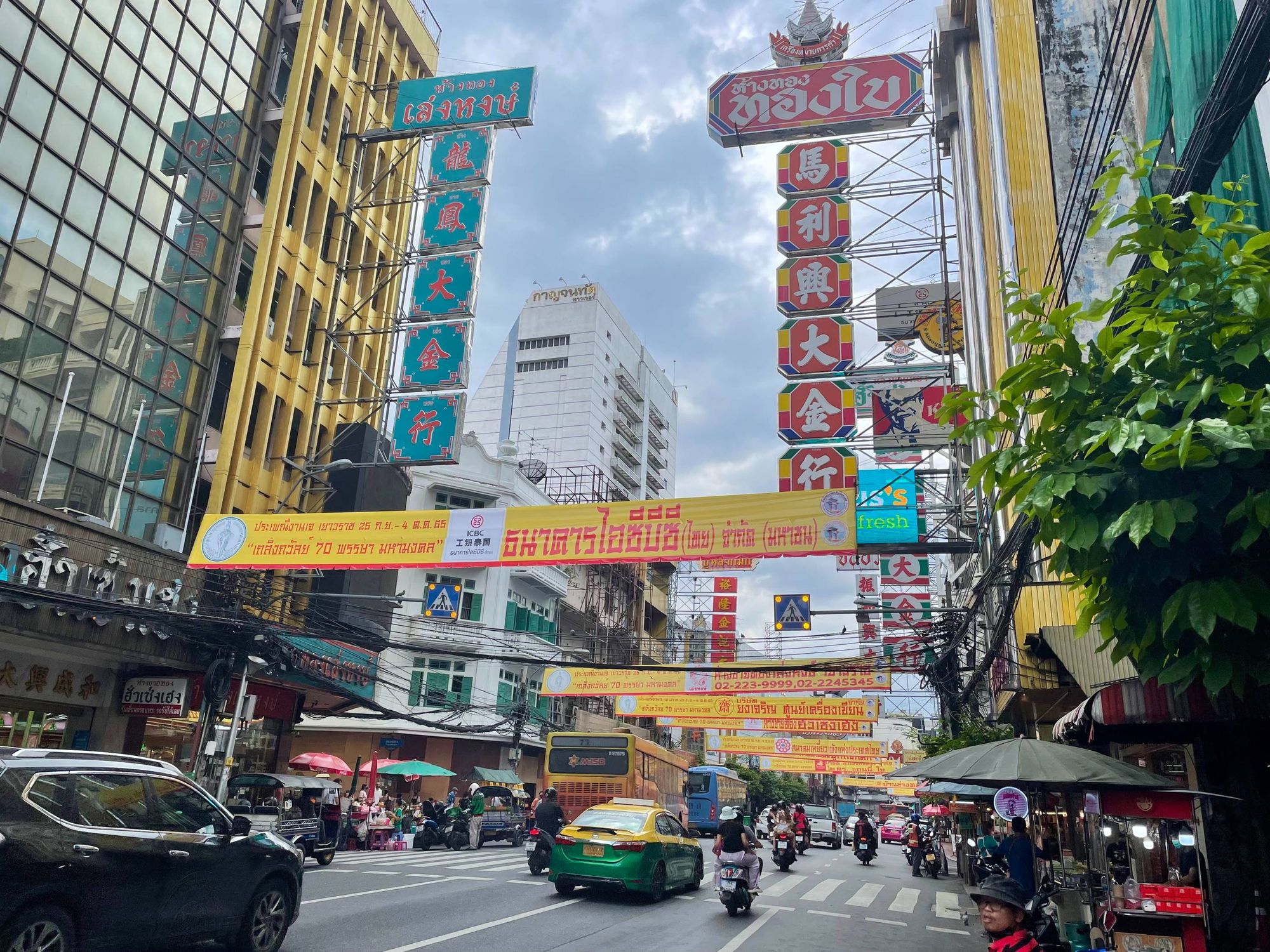 A busy street in Chinatown, Bangkok, Thailand. Lots of Chinese letters and billboards. There are cars, a yellow-green taxi and two buses. People riding on a scooter.