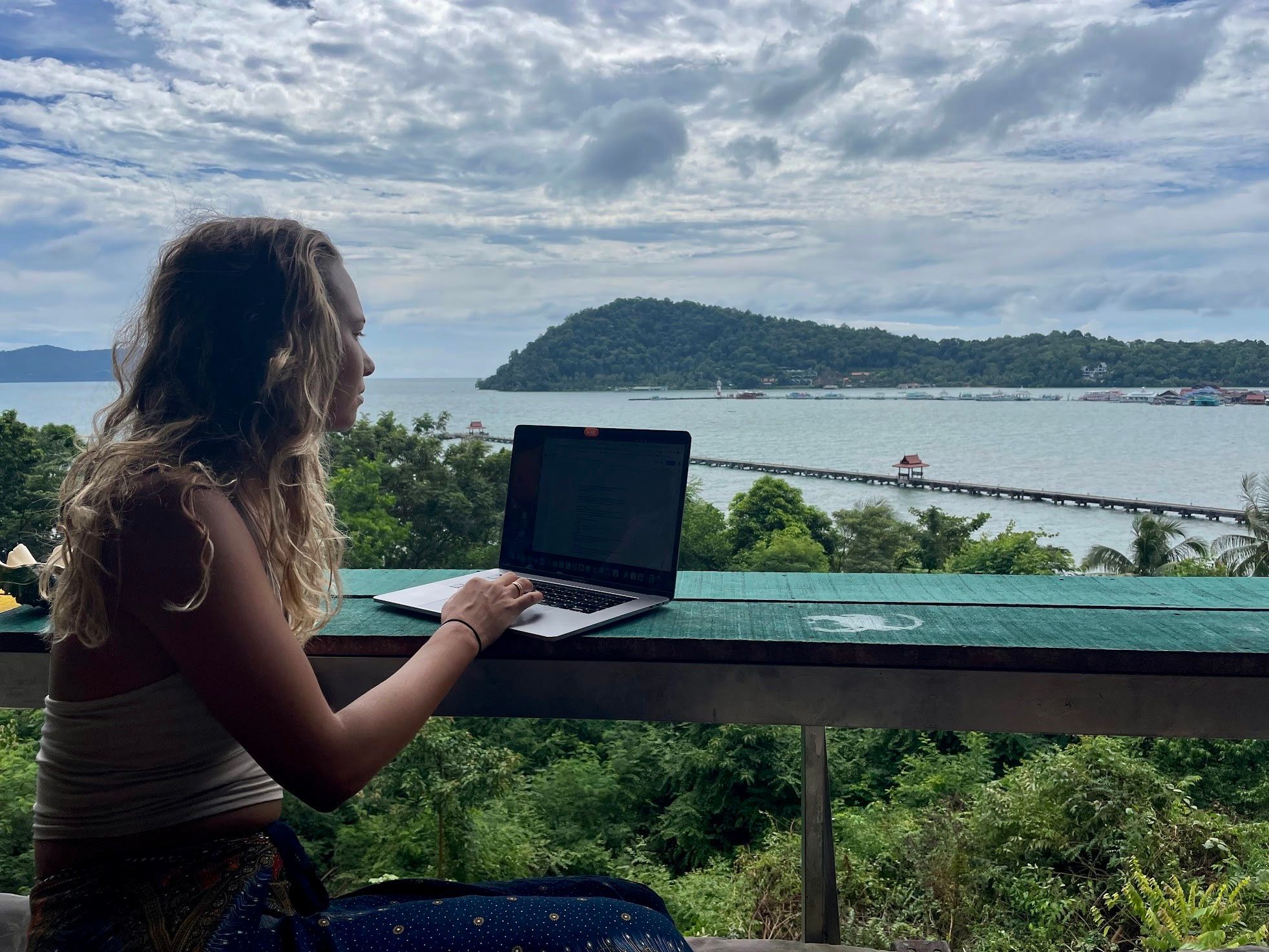 Lau working on her laptop with the sea and mountain in the background, lots of greenery. Located in Koh Chang, Thailand.