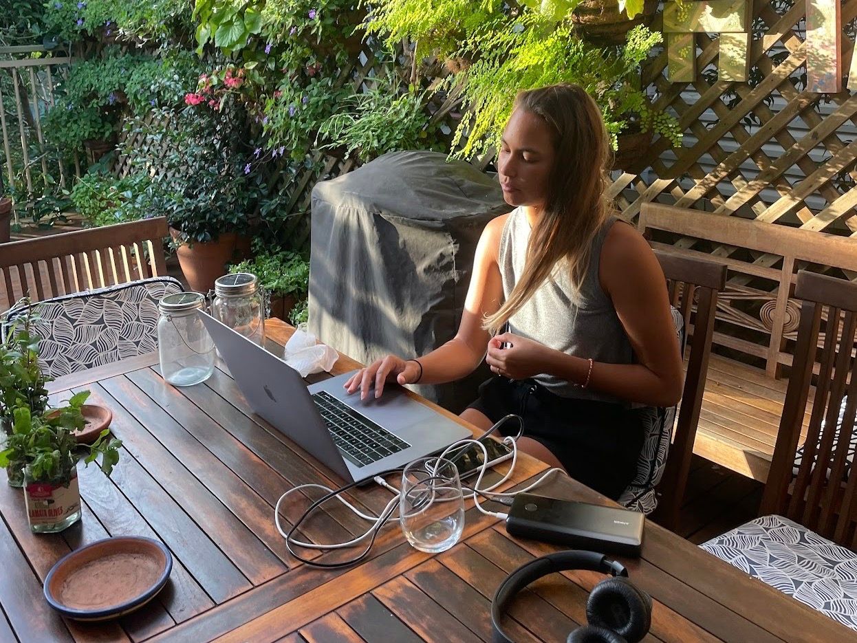 Laura working on her laptop in the garden, wooden table, chairs, plants in the background, headphones, glass of water, powerbank, phone