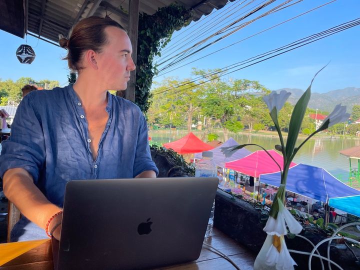 Izaac working on is MacBook laptop from a café in Mae Hong Son, Thailand. Flower on the table, riverview and markets